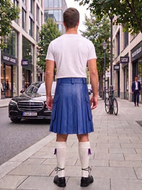 A man viewed from behind wearing a blue leather kilt, white t-shirt, white knee-high socks, and black shoes, standing on a city sidewalk.