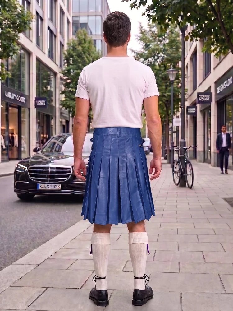 A man standing on a city sidewalk, viewed from behind, wearing a white t-shirt, a blue pleated leather kilt, white knee-high socks, and black shoes. Buildings, trees, a parked black car, and a bicycle are visible in the background.