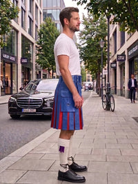 A man standing on a city sidewalk wearing a blue and red pleated kilt, a white t-shirt, white socks, and black leather shoes, with a street scene in the background.