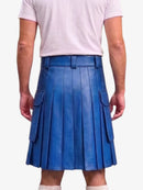 A man wearing a bright blue pleated faux-leather kilt with side cargo pockets, paired with a light pink t-shirt, shown from the back against a light gray background.