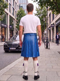 A man viewed from behind wearing a blue leather kilt, white t-shirt, white knee-high socks, and black shoes, standing on a city sidewalk.
