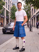 A man stands on a city sidewalk wearing a bright blue leather kilt, a white t-shirt, white knee-high socks, and black leather shoes. The background shows a street with buildings, a parked black Mercedes, and a bicycle.