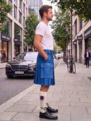 A man standing on a city sidewalk wearing a blue leather kilt, white t-shirt, white knee-high socks with a purple bowtie detail, and black leather shoes. A black Mercedes-Benz car and a bicycle are parked on the street behind him.