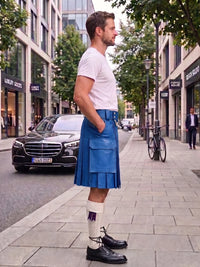 A man standing on a city sidewalk wearing a blue leather kilt, white t-shirt, white knee-high socks with a purple bowtie detail, and black leather shoes. A black Mercedes-Benz car and a bicycle are parked on the street behind him.