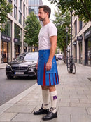 A man wearing a blue leather kilt with red and purple pleated accents, a white t-shirt, and white knee-high socks stands on a city sidewalk next to a street with cars and trees.