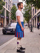 A man standing on a city sidewalk wearing a white t-shirt, a bright blue kilt with red pleats, white knee-high socks, and black leather shoes. The background shows a street with a black Mercedes-Benz car, storefronts, and a bicycle.