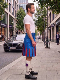 A man standing on a city sidewalk wearing a white t-shirt, a bright blue kilt with red pleats, white knee-high socks, and black leather shoes. The background shows a street with a black Mercedes-Benz car, storefronts, and a bicycle.