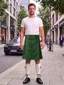 A man stands on a city sidewalk wearing a white crew-neck t-shirt and a green corduroy kilt with white knee-high socks and black leather shoes. A black Mercedes-Benz is parked in the background on a city street.