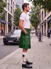 A man standing on a city sidewalk wearing a green leather kilt, white t-shirt, white socks, and black leather shoes, with a street scene in the background.