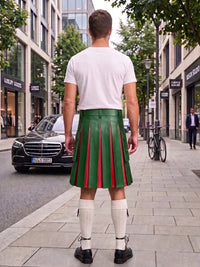 A man viewed from behind wearing a white t-shirt, a green leather pleated kilt with red pleats, white knee-high socks, and black shoes, standing on a city sidewalk.
