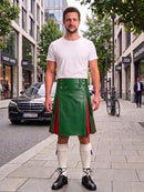 A man stands on a city sidewalk wearing a white t-shirt and a green leather kilt with red side panels, paired with white knee-high socks and black leather shoes. Buildings, trees, and a parked black car are visible in the background.