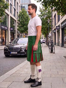 A man wearing a black leather kilt with red pleats, a white t-shirt, white knee-high socks, and black leather shoes stands on a city sidewalk next to a street with cars and trees.