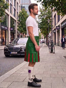 A man standing on a city sidewalk wearing a green kilt with red stripes, a white t-shirt, white socks, and black shoes. Buildings, trees, and parked cars are visible in the background.