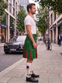 A man standing on a city sidewalk wearing a green kilt with red stripes, a white t-shirt, white socks, and black shoes. Buildings, trees, and parked cars are visible in the background.