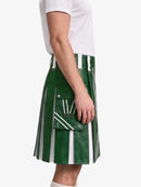 A side view of a person wearing a green leather kilt with white vertical stripes, white drawstrings, and a front utility pocket, paired with a white t-shirt against a light gray background.