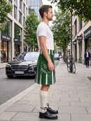A man standing on a city sidewalk wearing a white t-shirt, a green and white vertically striped kilt, cream knee-high socks, and black leather shoes. A black Mercedes-Benz car and a bicycle are visible in the background of the street scene.