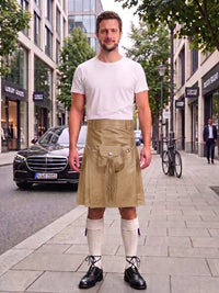 A man standing on a city sidewalk wearing a beige kilt with a front pouch pocket, a white t-shirt, white knee-high socks, and black leather shoes. Buildings, trees, and a black Mercedes-Benz car are visible in the background.