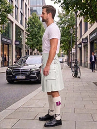 A man standing on a city sidewalk wearing a white t-shirt, a pale green pleated kilt with front pockets, white socks with purple accents, and black leather shoes. A black Mercedes-Benz car and urban buildings are visible in the background.