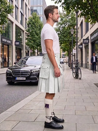 A man standing on a city sidewalk wearing a white short-sleeve T-shirt and a light gray pleated kilt with white socks and black shoes. Buildings, trees, a black Mercedes-Benz car, and a bicycle are visible in the urban street setting.