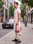 A man stands on a city sidewalk wearing a white pleated kilt with bold red and white vertical stripes, a white t-shirt, white socks, and black shoes. A black Mercedes-Benz car and a bicycle are parked on the street behind him, with trees and storefronts in the background.